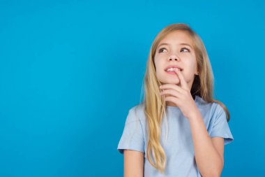  Düşünceli bir ifadeyle mavi tişört giyen güzel beyaz kız kameraya bakıyor, elini yüzüne yaklaştırıyor, parmağını ısırıyor hoş bir şey düşünüyor..