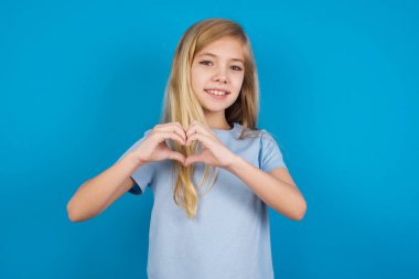  Güzel beyaz kız mavi tişört giyiyor. Sevgiyle gülümsüyor. Elleriyle kalp sembolü ve şekli gösteriyor. Romantik konsept.