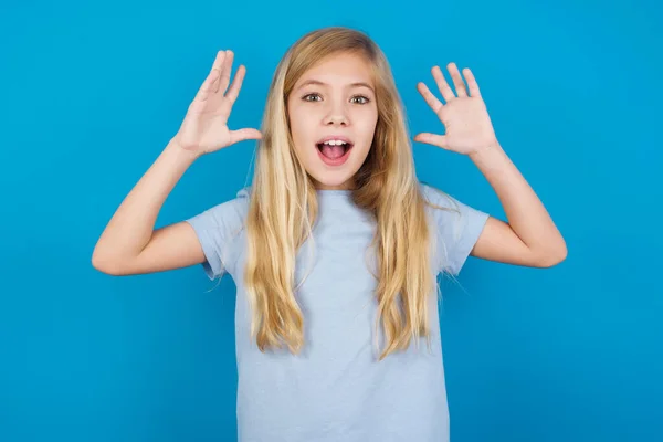 青いTシャツを着ている美しい白人少女はカメラで興奮して見て 頭の上に手を上げ 予期しないことに気づき 突然のニュースに反応します — ストック写真