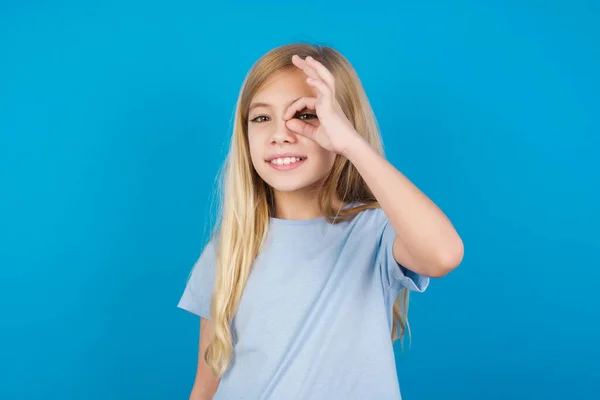 美しい白人少女は 幸せな顔で青いTシャツを着て 指を通して見て手でOkのサインをする — ストック写真