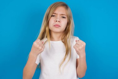 Displeased annoyed beautiful caucasian girl wearing t-shirt clenches fists, gestures pissed, ready to revenge, looks with aggression at camera stands full of hate, being pressured