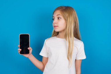  Tişört giyen güzel beyaz kız yeni bir cep telefonu tutuyor ve gizemli görünüyor. Modern cep telefonunun boş görüntülerini gösteriyor.