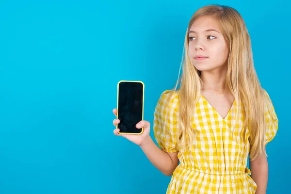ドレスを着ている美しい白人少女は新しいスマートフォンを保持し 現代の細胞の空白の表示を示す不思議な脇に見えます — ストック写真
