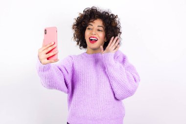  Süveter giyen genç ve güzel İspanyol kadın modern akıllı telefonu tutuyor ve merhaba jesti olarak el sallıyor. İnsanlar modern teknoloji kavramı