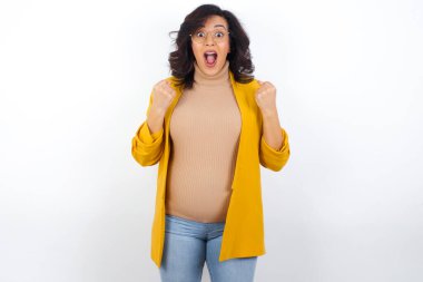  Sarı spor ceket giyen hamile bir kadın başarı ve zaferle sevinçle yumruklarını sıkıp amacına ve amaçlarına ulaştığına seviniyor. Olumlu duygular, duygular.