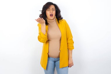 Studio shot of frustrated  pregnant woman wearing yellow blazer gesturing with raised palm, frowning, being displeased and confused with dumb question.