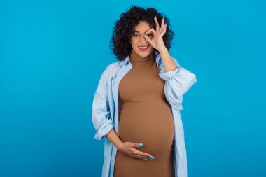  Hamile Arap kadın el hareketleriyle gülümsüyor, gözleriyle mutlu yüzüyle parmaklarının arasından bakıyor..