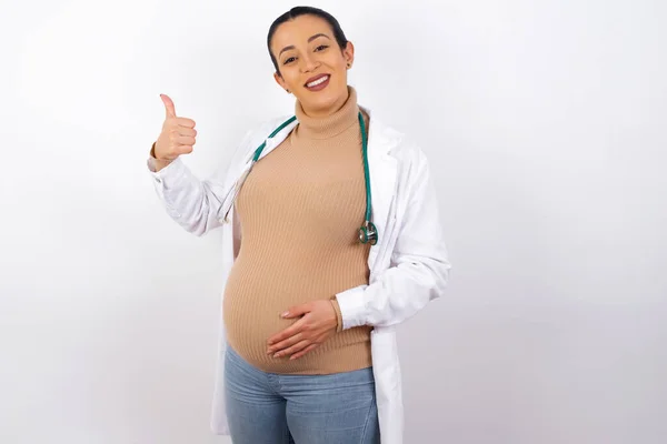 年轻而漂亮的怀孕医生 身穿医疗制服 身披大拇指 背景偏僻 — 图库照片