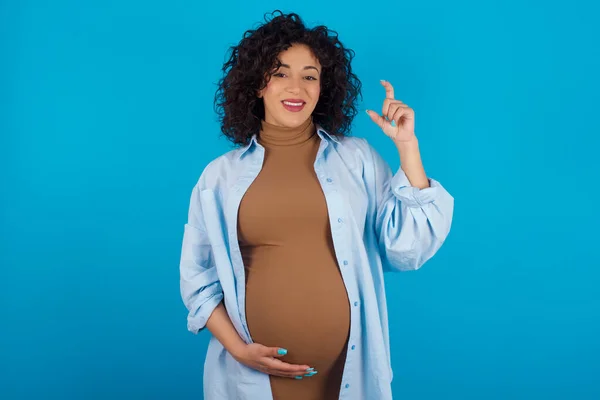 妊娠中のアラブの女性は笑顔で自信を持って指を見て小さなサイズのサインをする手でジェスチャーします 測定コンセプト — ストック写真