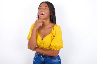 Sarı bluz ve kot pantolon giyen genç ve güzel bir kadın mutlu bir şekilde gülüyor. Elini çenesinde tutuyor. Olumlu duygular ifade ediyor. 