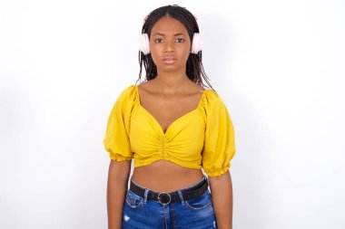 Ciddi şekilde hoşnutsuz, sarı bluz giyen ve kot giyen genç bir kadın kameraya şaşırmış görünüyor kızgın olmak müzik kulaklığı takıyor müzik dinliyor. 