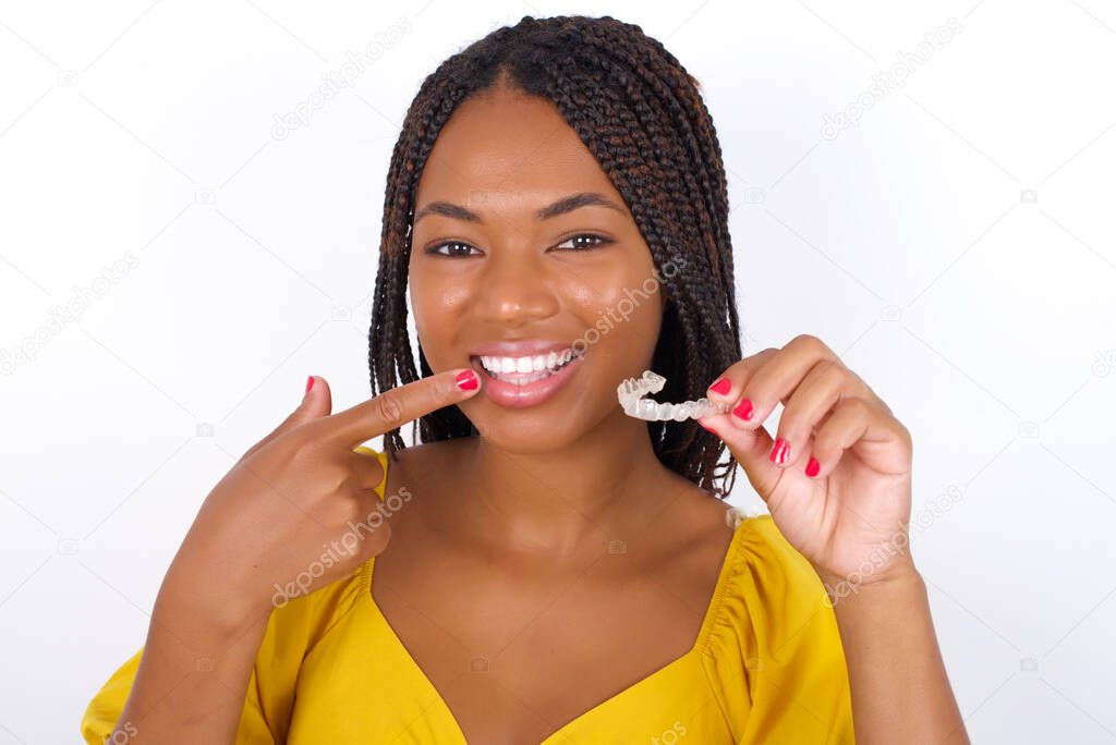 young beautiful woman wearing yelllow top and jeans   holding an invisible aligner and pointing to her perfect straight teeth. Dental healthcare and confidence concept.