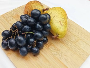 Minimalist meyve, iki olgun armut ve bambu tahtasına yerleştirilmiş büyük bir demet siyah üzüm içerir. Doğal sıcak renkler ve temiz kompozisyon, sağlıklı yemek temaları için mükemmel bir besin tasarımı tarifi sağlar. 