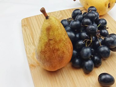 Olgun armut ve temiz beyaz arka planlı tahta bir kesme tahtasına dizilmiş bir demet siyah üzüm. Sağlıklı yemek temaları, organik ürünler, mevsimsel yiyecekler, beslenme ve minimal gıda fotoğrafçılığı için doğal meyve bileşimi.