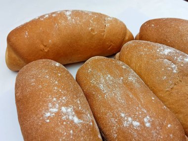 Temiz beyaz bir zemin üzerine hafifçe un serpiştirilmiş taze altın fırınlı ekmek rulolarını kapat. Yumuşak doku, ev yapımı kırsal yapı ve sıcak pişirme atmosferi. Fırın menüleri, gıda ambalajları, tarif blogları, yemek temaları ve organik