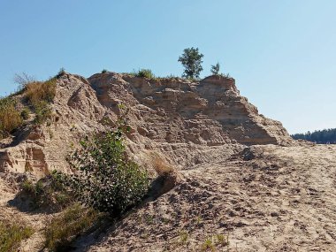 Kumlu taş ocağı manzarası katmanlı toprak, erozyon desenleri ve açık mavi gökyüzünün altında seyrek bitki örtüsü. Madencilik ve çevresel süreçler tarafından şekillendirilen doğal arazi. Jeoloji kavramı, arazi bozulması, taş ocağı, çevresel etki ve peyzaj Chan