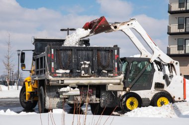 Büyük metal bir kazıcı kovası şehir yolundan bir kamyona kar yüklüyor.