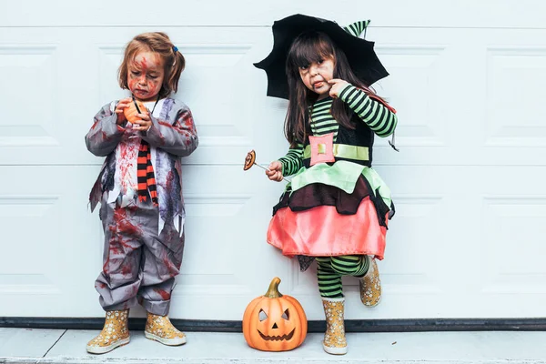 Cadılar Bayramı 'nı Jack O Lantern' in yanındaki garaj kapısında şeker yiyen cadı ve zombi gibi giyinmiş iki kız.