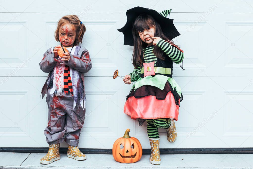 Two girls dressed as witch and zombie eating candy celebrating Halloween at the garage door next to Jack O Lantern