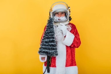 Astronot kasklı Noel Baba sarı arka planda bir Noel ağacı tutuyor. Noel konsepti