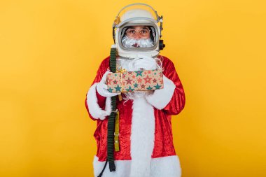 Astronot kasklı Noel Baba sarı arka planda bir hediye tutuyor. Noel konsepti