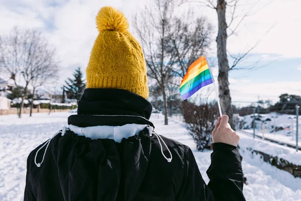 Lesbian woman with her back turned wearing a yellow snow cap, in a snowy area holding lgbt rainbow flag
