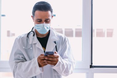 Beyaz önlüklü, steteskop takmış siyah saçlı bir doktor, vitrindeki cep telefonuna bakıyor. Tıp konsepti