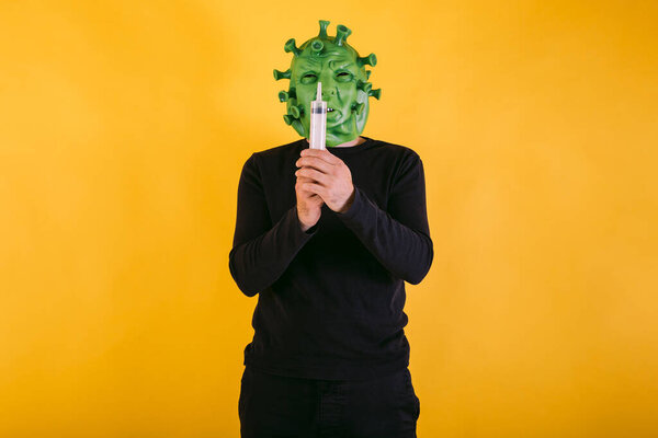 Человек, замаскированный под коронавирус с латексной маской - вирус ковид-19, держащий шприц на желтом фоне. Коронавирусная концепция