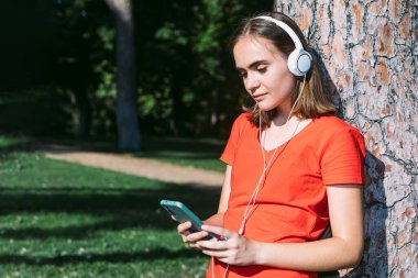 Kırmızı tişörtlü genç bir sarışın cep telefonuna bakar ve bir parkta müzik dinler. Ağacın gövdesine yaslanır.