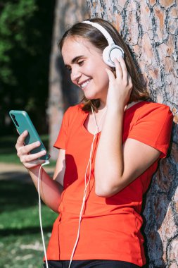 Kırmızı tişörtlü genç bir sarışın cep telefonuna bakar ve bir parkta müzik dinler. Ağacın gövdesine yaslanır.