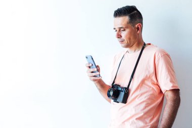 Omzunda eski moda fotoğraf makinesi olan pembe tişörtlü bir adam cep telefonuna bakıyor.