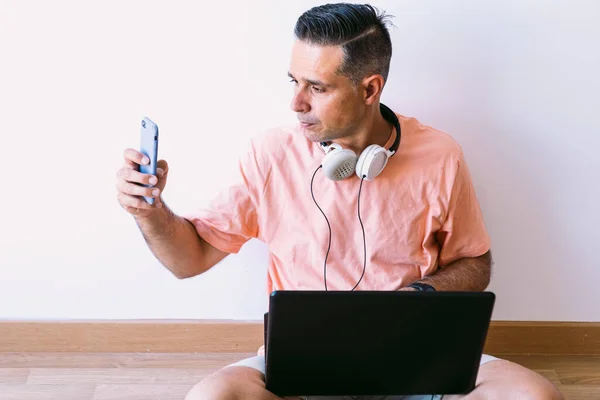 Man sitting on the floor with his laptop teleworking from home, wearing headphones and looking at the mobile phone