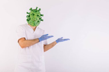 Koronavirüs kılığına girip lateks maske takan, doktor kıyafeti giyen ve beyaz arka planda iki eliyle sağı gösteren kişi. Coronavirus konsepti