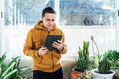 Pencerenin kenarında yeşil çiçekler ve bitkilerle çevrili bir tablete bakan bir adam.
