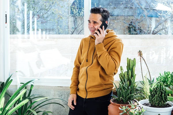 Smiling man talking on the mobile near the window, surrounded by green plants and flowers