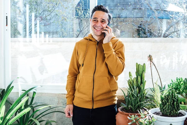 Smiling man talking on the mobile near the window, surrounded by green plants and flowers