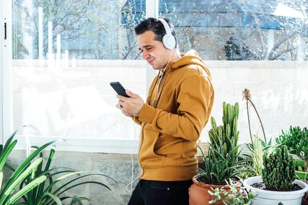 Smiling man looking at the mobile and listening to music with headphones near the window, surrounded by green plants and flowers