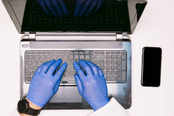 Aerial view of a laptop, doctor\'s hands with latex gloves typing with the keyboard, with a smartphone on the right