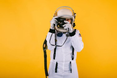 Uzay giysisi ve kasklı bir erkek kozmonot, sarı arka planda retro kamerayla fotoğraf çekiyor..