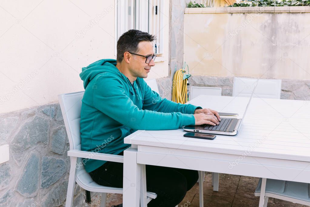 Young man telecommuting, typing on his laptop in the garden of his house