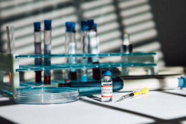 Test tüpleri için tutacağı olan bir laboratuvar masası COVID-19 aşısı olan bir şişe şırınga, pipet ve petri kabı olan bir perde gölgesi olan bir şişe.