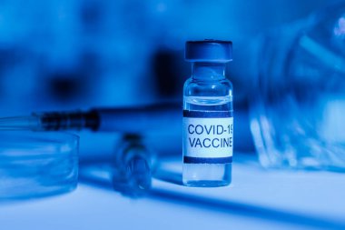 Test tüpleri destekli laboratuvar masası, COVID-19 aşısı şişesi, şırınga, pipet, koruyucu gözlük ve petri kabı, mavi ışık ortamı