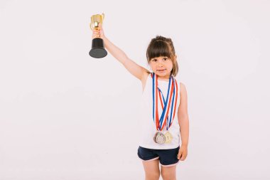 Spor şampiyonluğu madalyaları olan kahverengi saçlı küçük kız tek eliyle kupayı kaldırıyor. Spor ve zafer kavramı