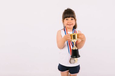 Spor şampiyonluğu madalyaları olan kahverengi saçlı küçük bir kız, iki eliyle bir kupa tutuyor. Spor ve zafer kavramı