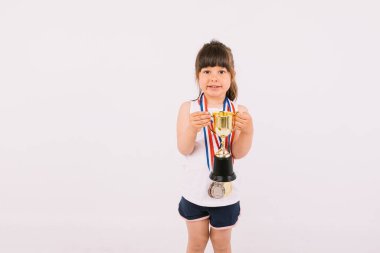 Spor şampiyonluğu madalyaları olan kahverengi saçlı küçük bir kız, iki eliyle bir kupa tutuyor. Spor ve zafer kavramı