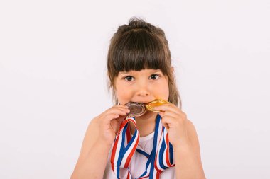 Spor şampiyonluğu madalyaları olan kahverengi saçlı küçük kız onları ısırıyor. Spor ve zafer kavramı