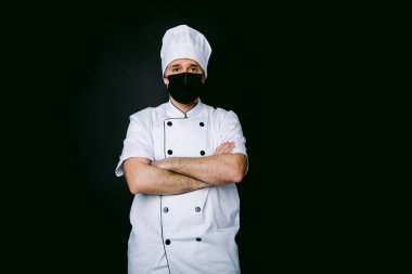 Aşçı mutfak ceketi ve şapka giyiyor, siyah maskeli, kolları çapraz, siyah arka planda.
