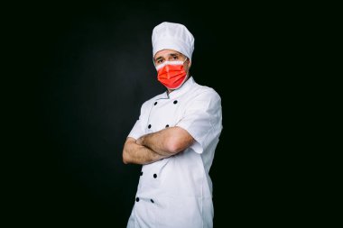 Aşçı kırmızı maskeli, yemek pişirme ceketi ve şapkası giymiş, kollarını kavuşturmuş, siyah arka planda.