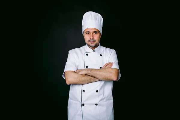 Aşçı yemek pişirme ceketi ve şapka giymiş, kollarını kavuşturmuş, siyah arka planda.