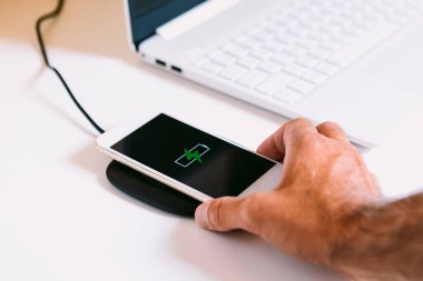 Ekranında yeşil yıldırım olan beyaz cep telefonu logosuyla el, beyaz çalışma masasındaki dizüstü bilgisayarın yanındaki kablosuz şarj tabanında şarj oluyor.
